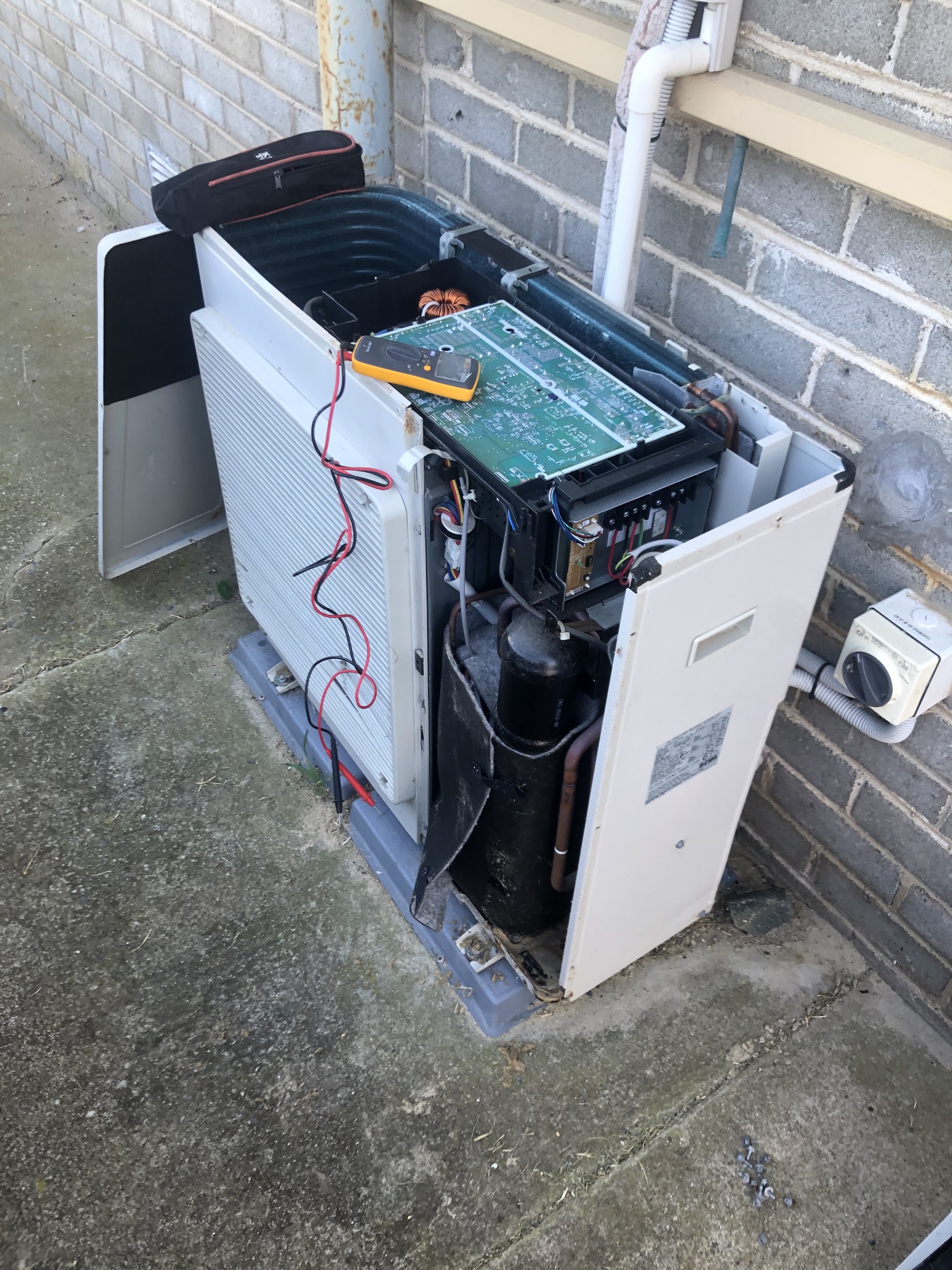 An outdoor air conditioning unit stripped to the chassis with its control board lifted out and a multimeter across the terminals